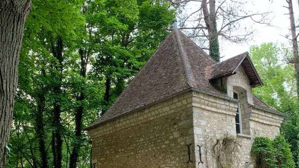 Edifício de pedra com telhado de telhas, rodeado por árvores. Há mesas e cadeiras ao ar livre, e um cão é visível.
