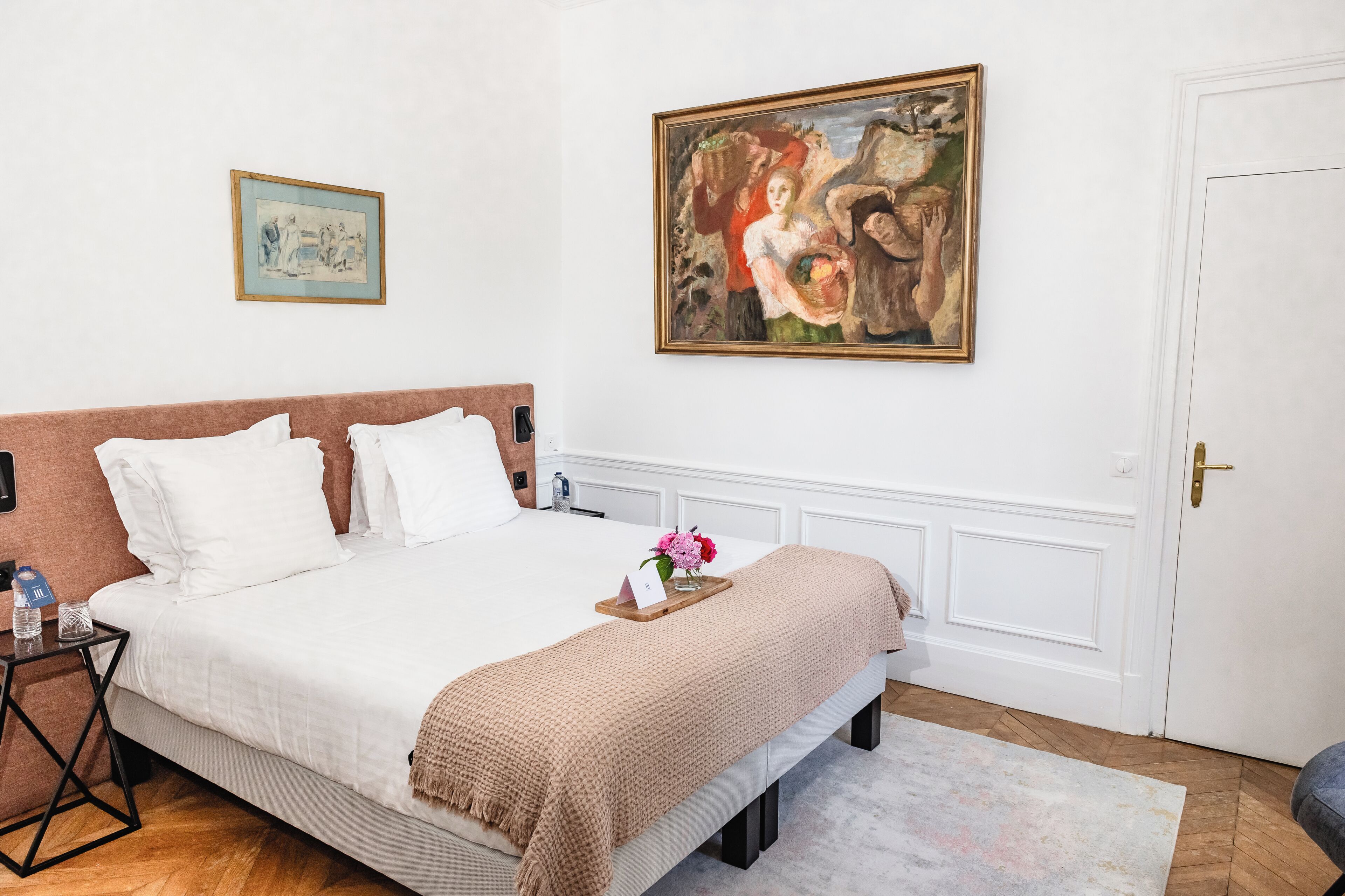 A neatly made bed with white linens, a brown throw blanket, and a small bedside table with a water bottle and a vase with flowers.