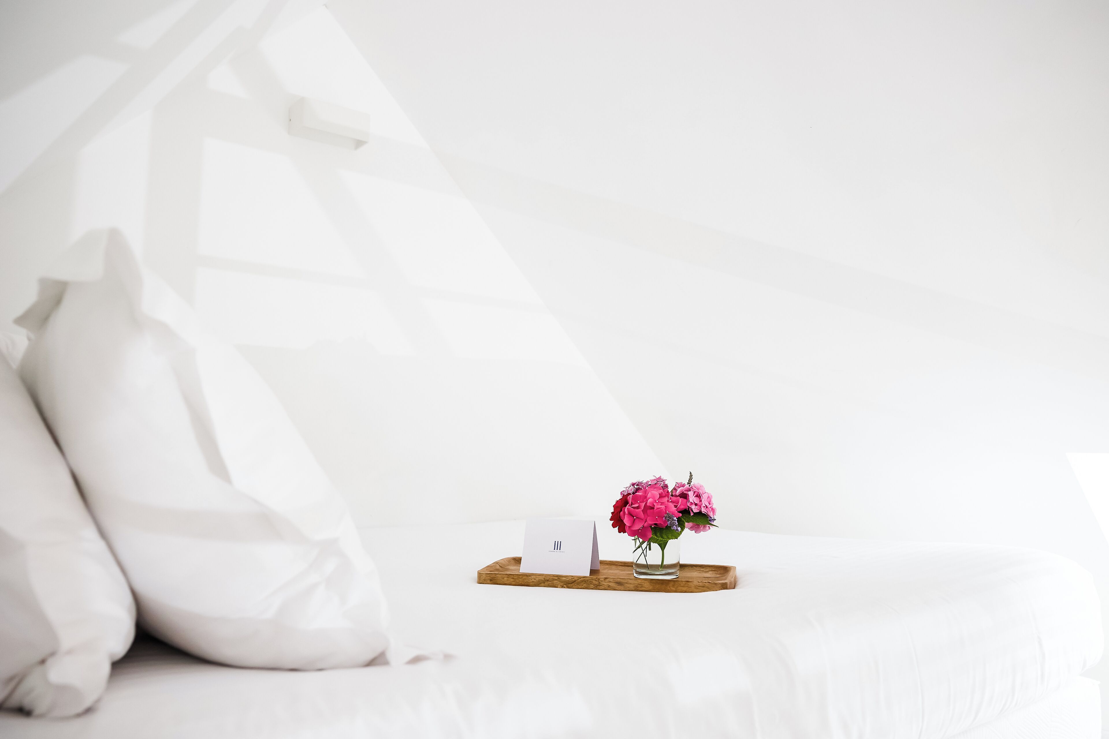 Une pièce blanche avec un lit, un plateau en bois sur lequel se trouve un vase de fleurs et une petite carte.