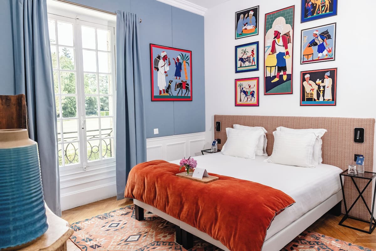 Quarto de hotel com uma cama grande, janela com cortinas, parede com obras de arte emolduradas e uma pequena mesa de cabeceira.