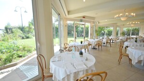 A restaurant with round tables and wicker chairs, large windows offering a view of greenery. - Bellevue Beach (Cervia)