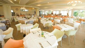A spacious dining area with round tables and chairs, a kitchen in the background, and large windows providing natural light. - Bellevue Beach (Cervia)