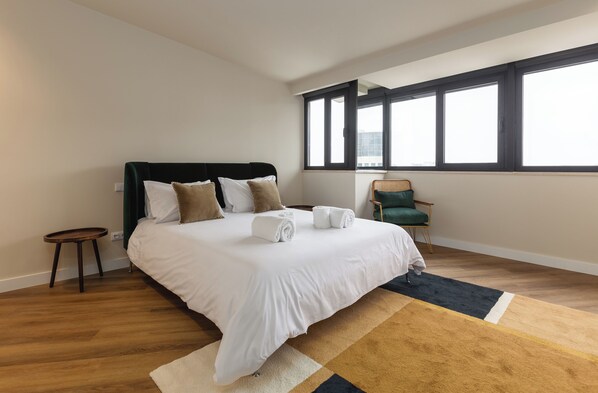 A modern hotel room with a large bed, a small side table, a chair, and a view of the city through large windows.