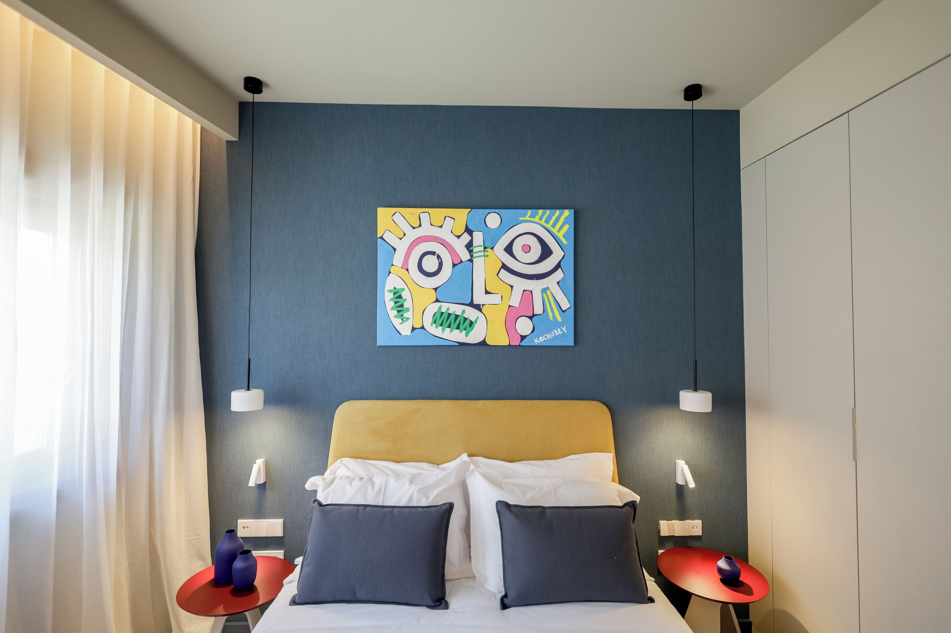 Moderni hotellihuone, jossa on sänky, kaksi yöpöytää, seinälle ripustettu taulu ja ikkuna, jossa on verhot.