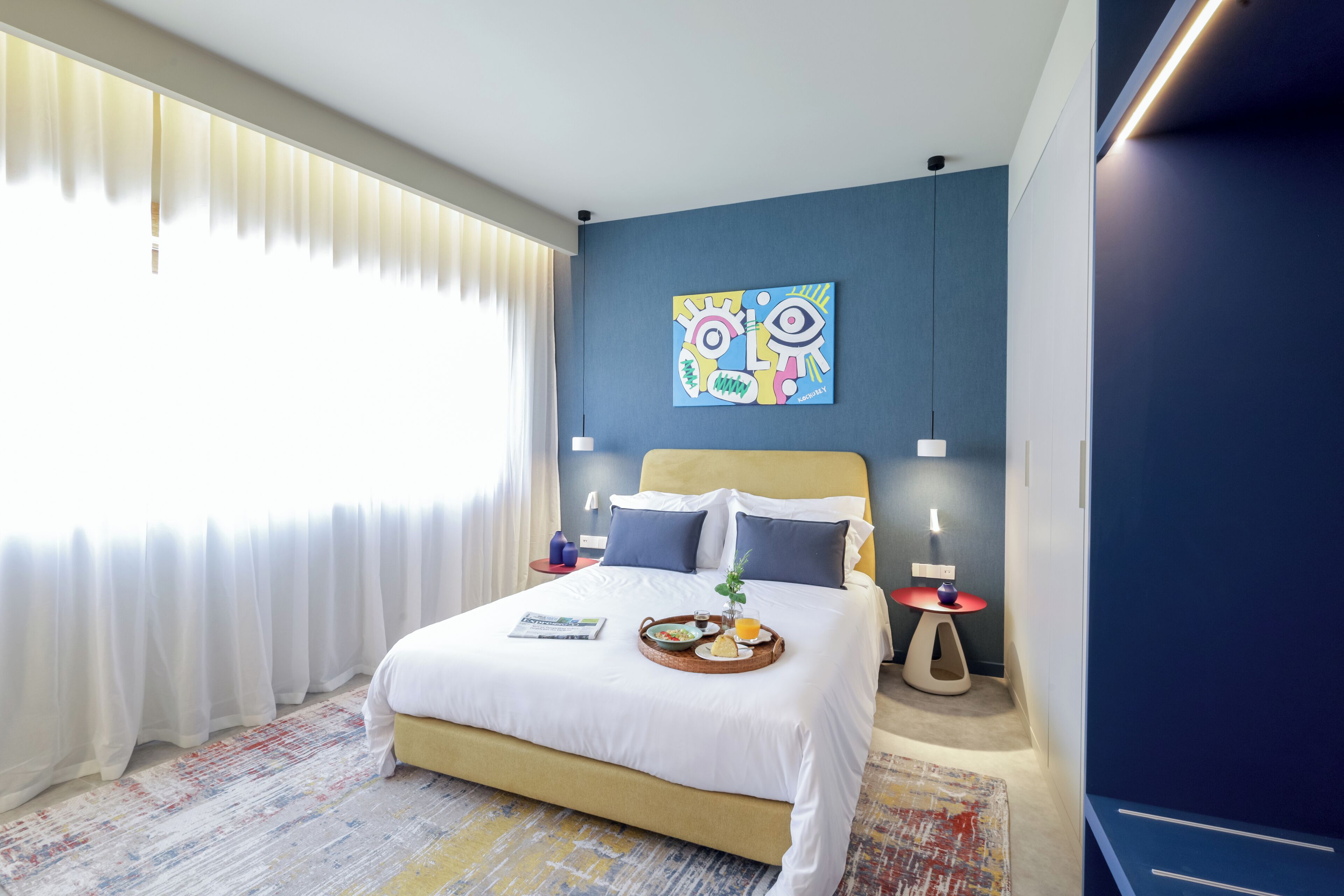 A hotel room with a bed, a painting on the wall, a tray with breakfast, and a window with curtains.