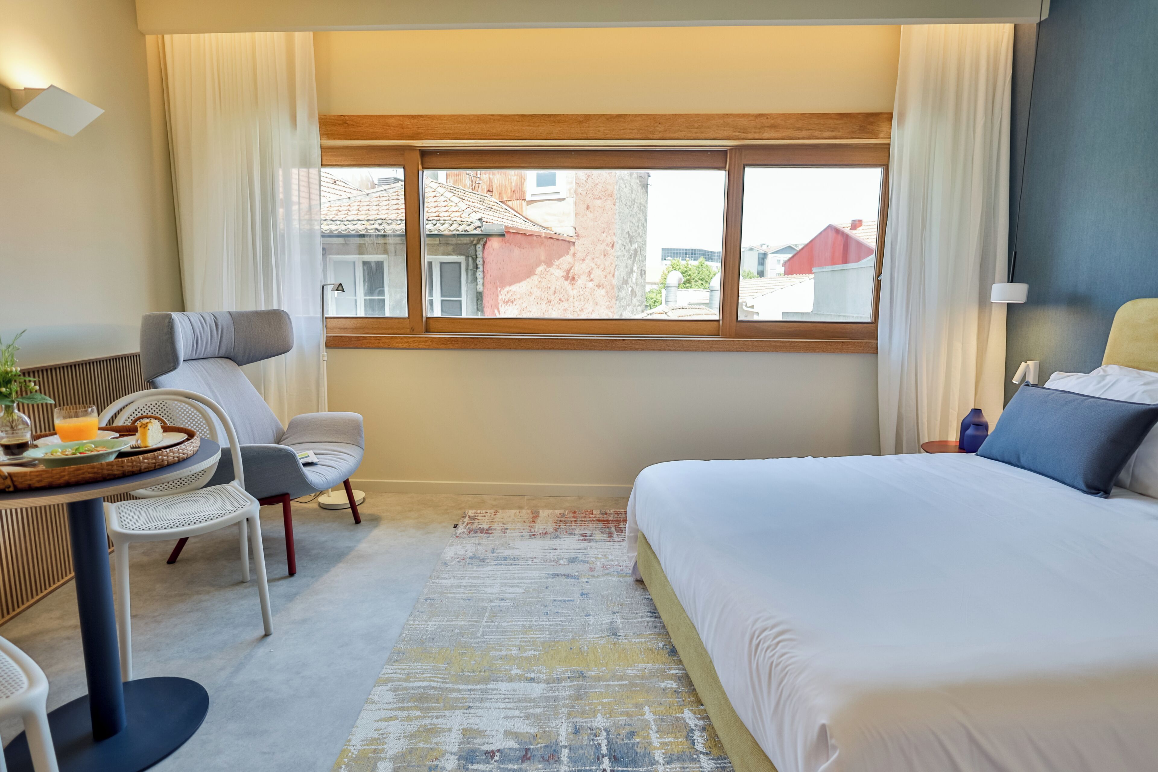 Habitación de hotel con cama, un escritorio con silla, una pequeña mesa con provisiones para el desayuno y vistas de edificios a través de la ventana.