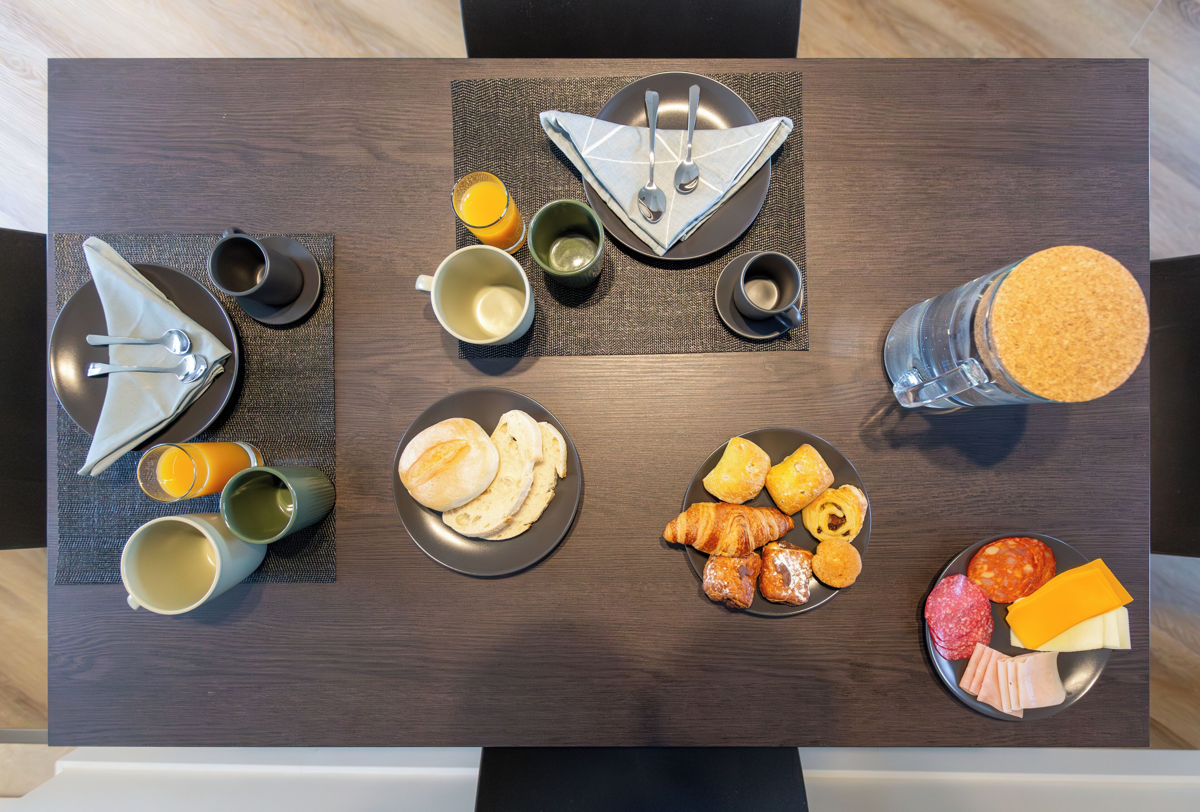 Una mesa de desayuno bien dispuesta con platos de pasteles, queso y embutidos, acompañada de tazas y un vaso de jugo de naranja.