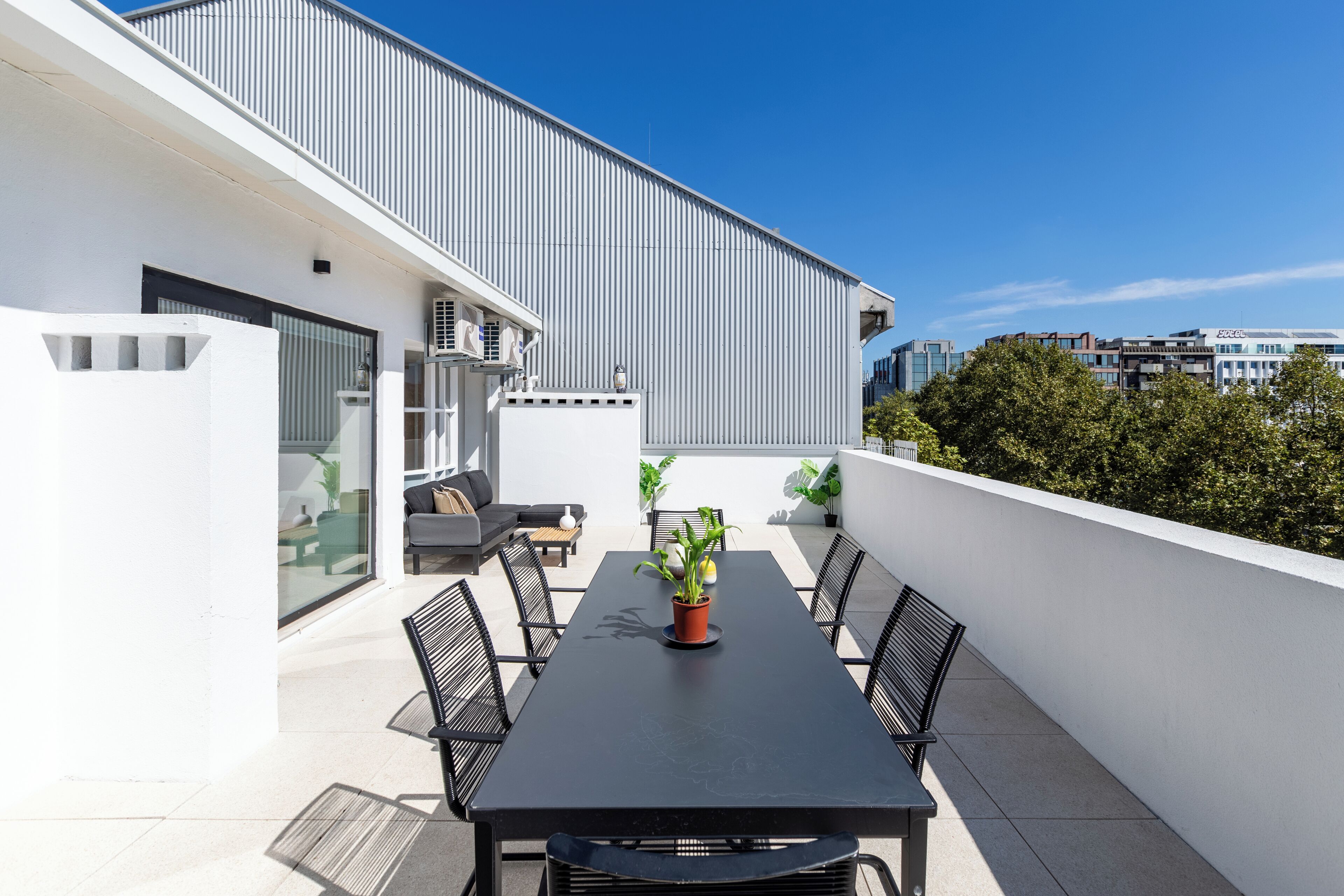 A modern outdoor patio with a black table and chairs, a potted plant, and a view of a building with a white facade and a sloped roof.