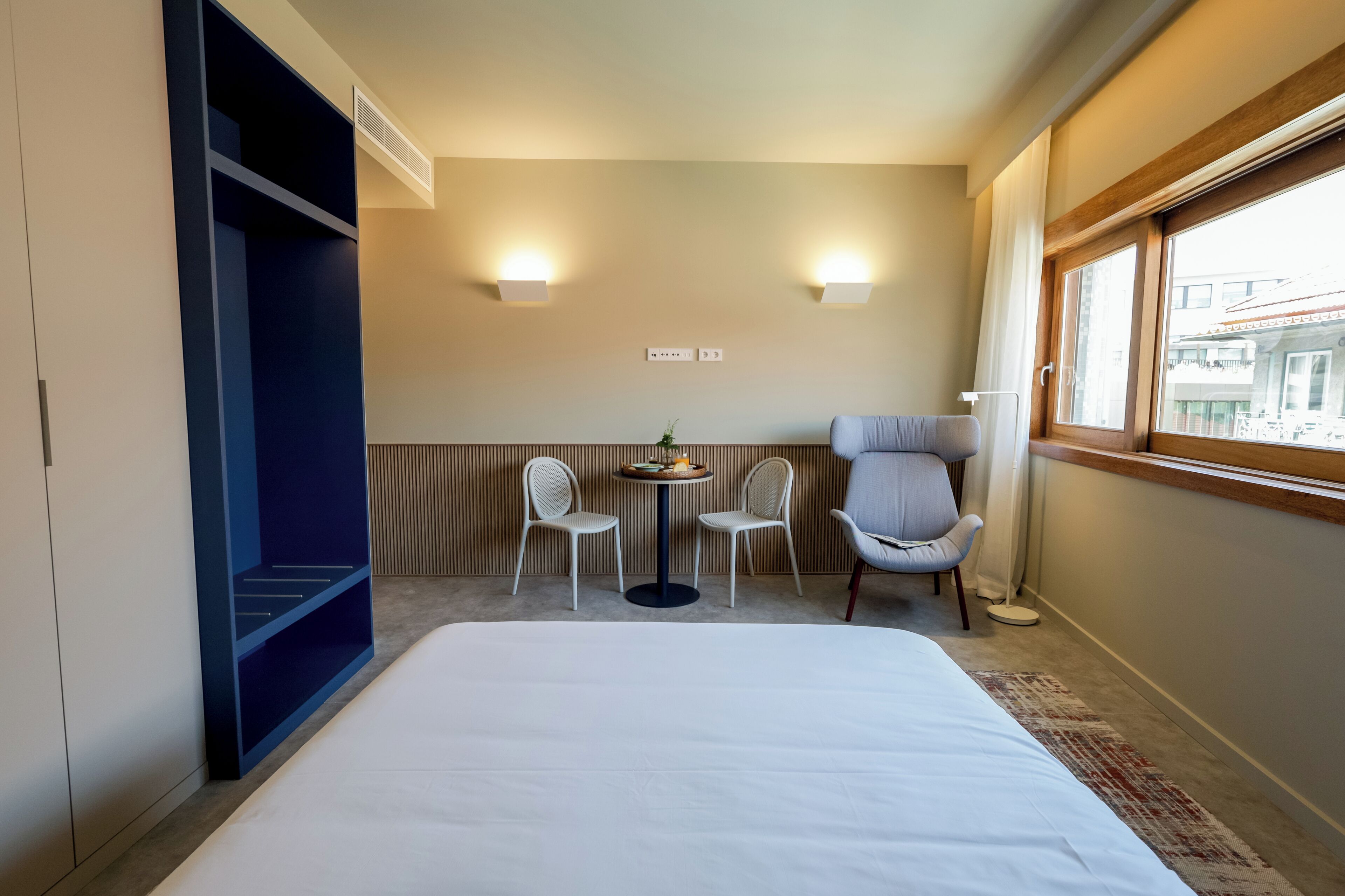A hotel room with a bed, a desk with two chairs, a small table with a plant, and a window with a view of buildings.