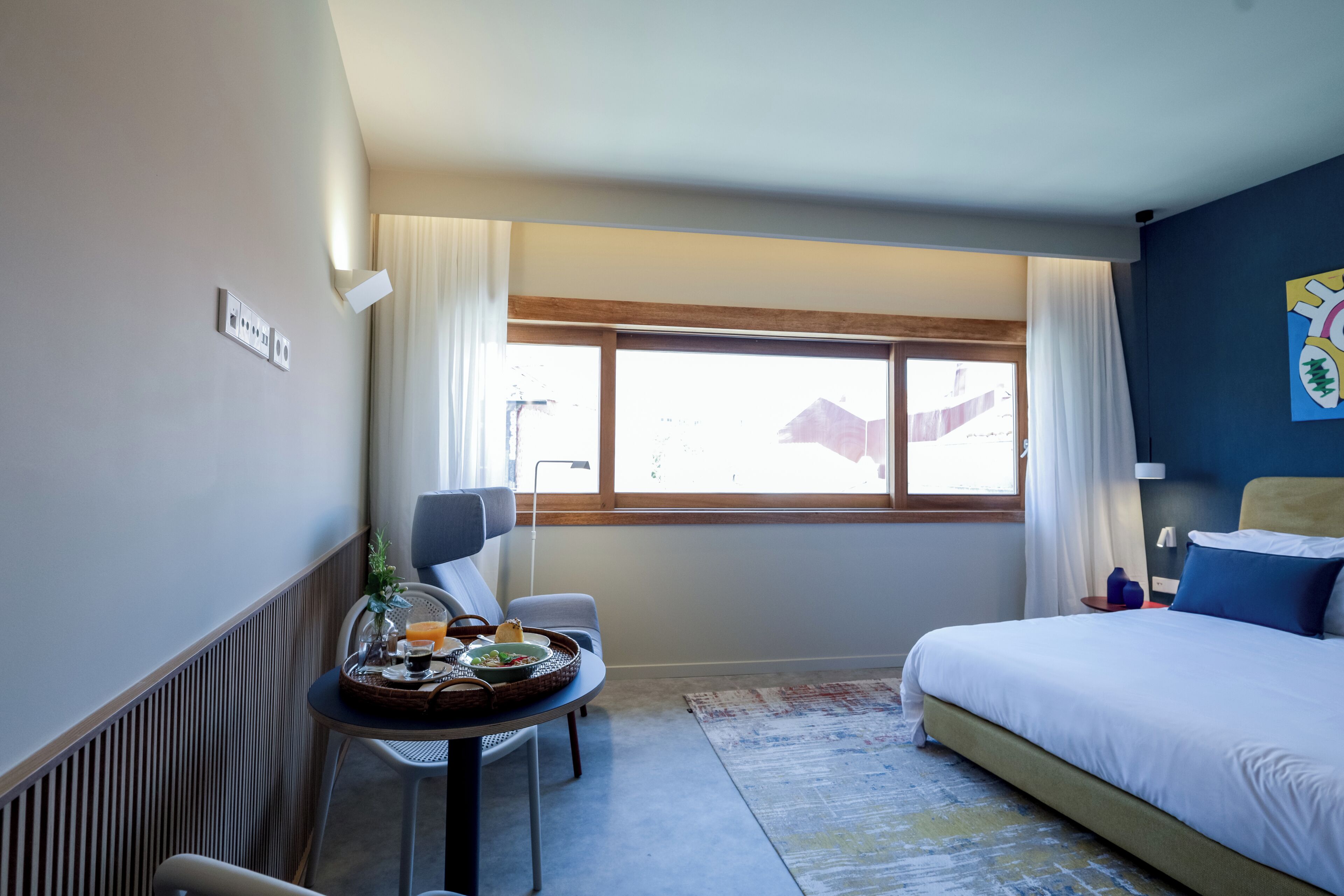 A hotel room with a bed, a small table with breakfast items, a chair, a window with curtains, and a view of the outside.