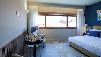 A hotel room with a bed, a small table with breakfast items, a chair, a window with curtains, and a view of the outside.