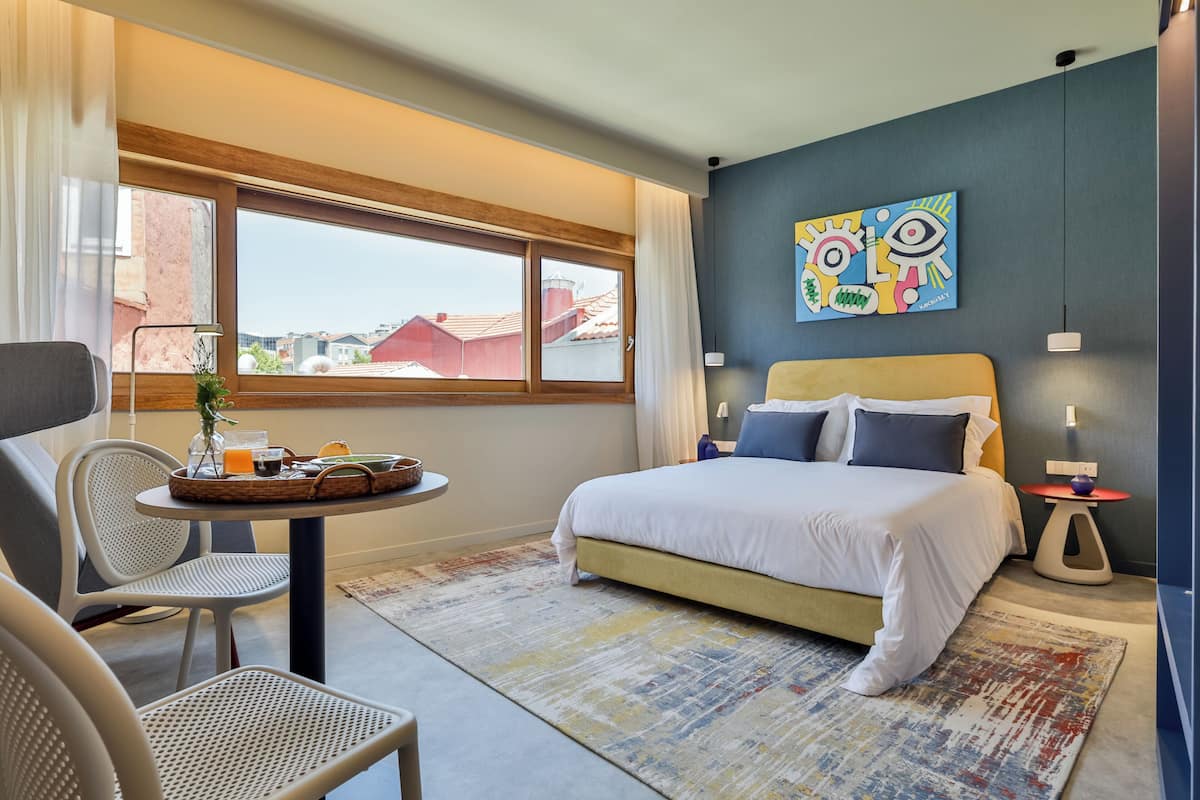 Habitación de hotel moderna con una cama grande, un área de comedor con mesa y sillas, y vistas de edificios a través de la ventana.