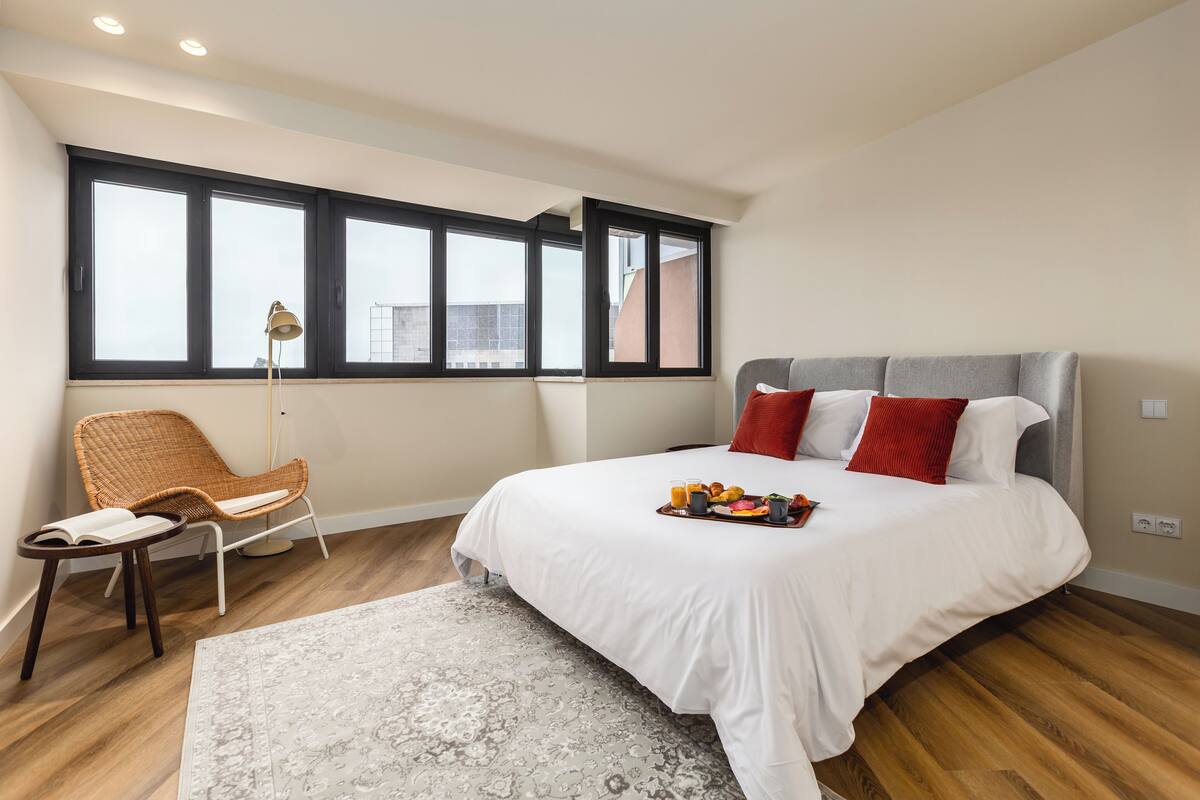 Un dormitorio moderno con una cama grande, una silla de mimbre, una lámpara de pie y vistas a la ciudad a través de amplias ventanas.