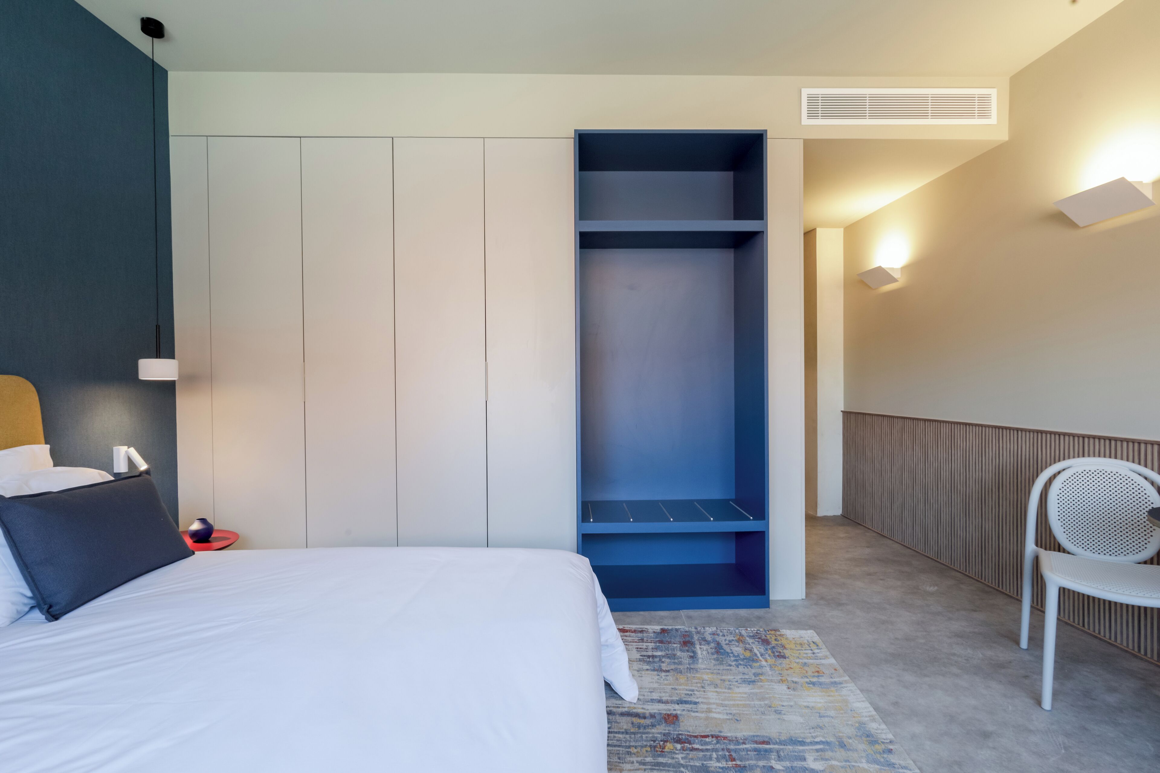 Une chambre d’hôtel moderne équipée d’un lit, d’une armoire intégrée, d’un coin salon et d’un luminaire fixé au mur.