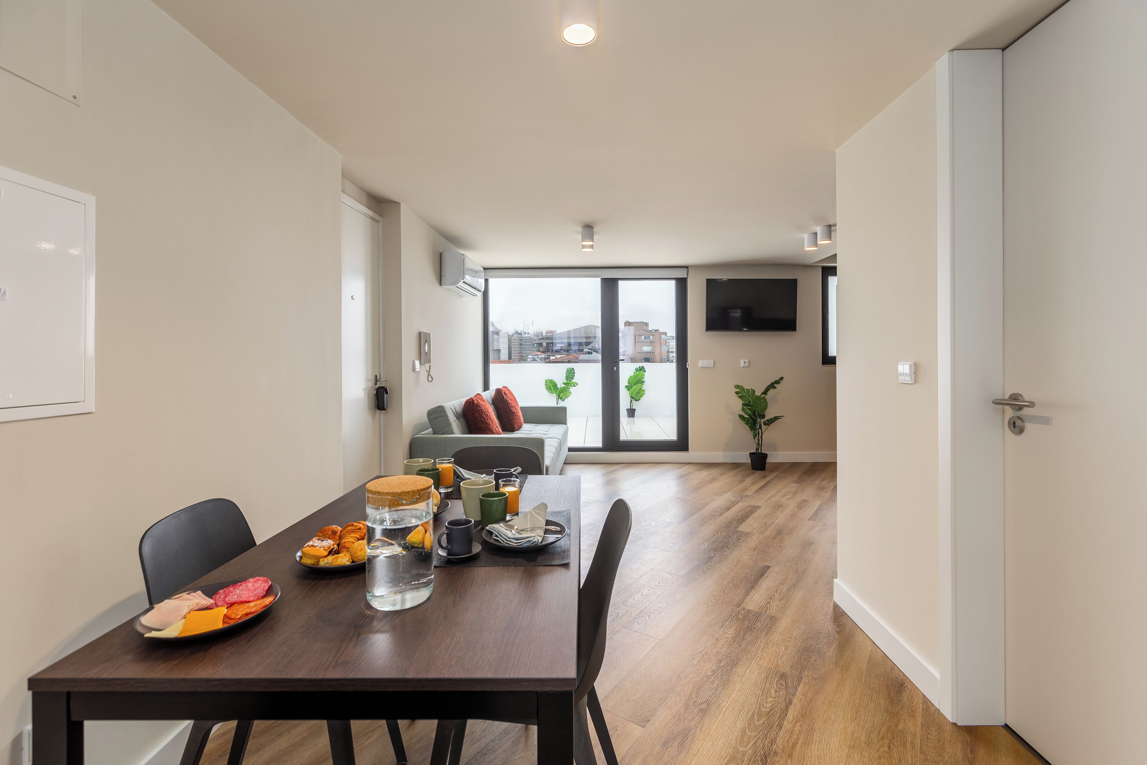 A modern dining area with a wooden table set for breakfast, a sofa with cushions, and a view of the city through large glass doors.
