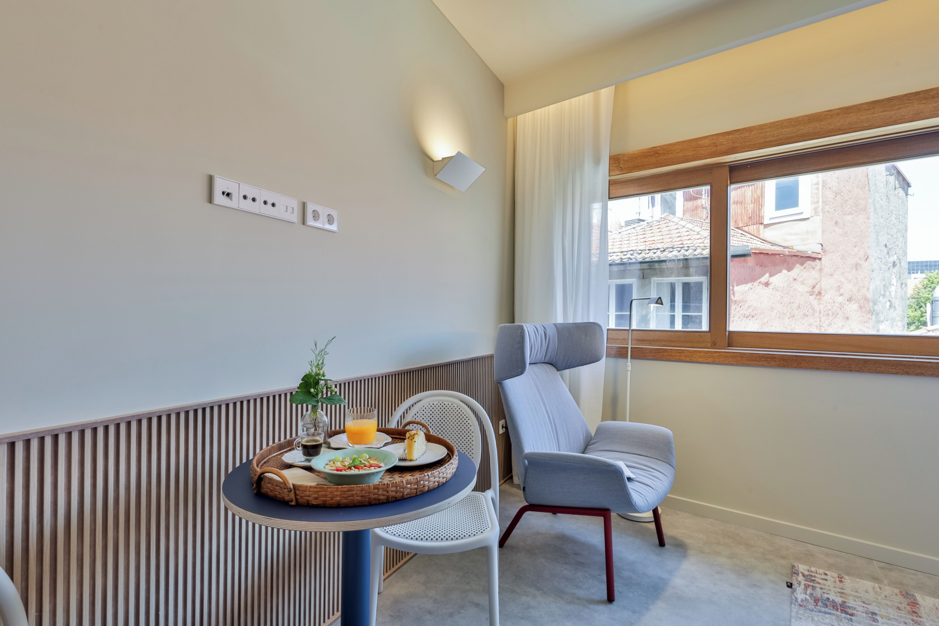 A modern room with a dining area featuring a round table with a basket, a chair, and a small potted plant. There is a window with a view of buildings outside.