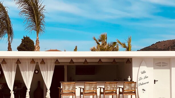 A poolside area with a table and chairs, a white wall with a surfboard, and palm trees. - Villa Las Palmeras in Alhaurin de la Torre (Alhaurin de la Torre)