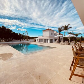A modern pool area with a clear sky, a pool, lounge chairs, and a building with a red roof. - Villa Las Palmeras in Alhaurin de la Torre (Alhaurin de la Torre)