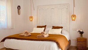 A neatly made bed with a brown and white bedspread, two pillows, and two rolled towels. There are two hanging lamps with a woven design, a white door with paneling, and a small round wooden table with a vase of flowers. - Villa Las Palmeras in Alhaurin de la Torre (Alhaurin de la Torre)
