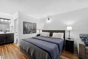 A bedroom with a bed, bedside lamps, a nightstand, a chair, a painting, and a bathroom with a mirror and sink. - Condo in the Heart of Shanty Creek Resort! (Bellaire)