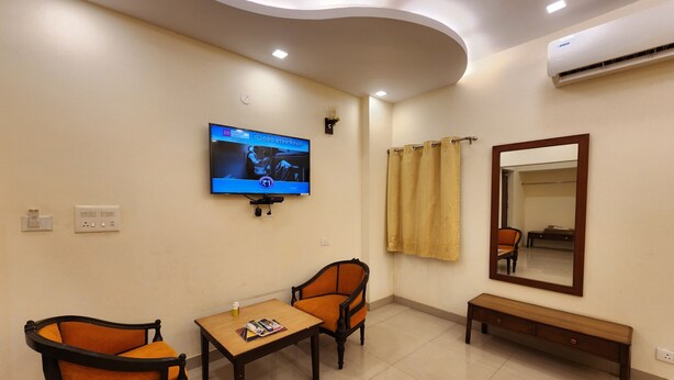 A hotel room with a television mounted on the wall, a bench, two chairs, a table, and a mirror.