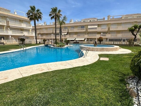A swimming pool surrounded by a well-maintained lawn, palm trees, and a multi-story hotel building with balconies. - Beautiful apartment, very well-equipped, air conditioning, Wi-Fi, satellite TV. (Alacant)