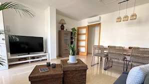 A modern living room with a flat-screen TV, wicker furniture, a dining area with wicker chairs, and a large potted plant.