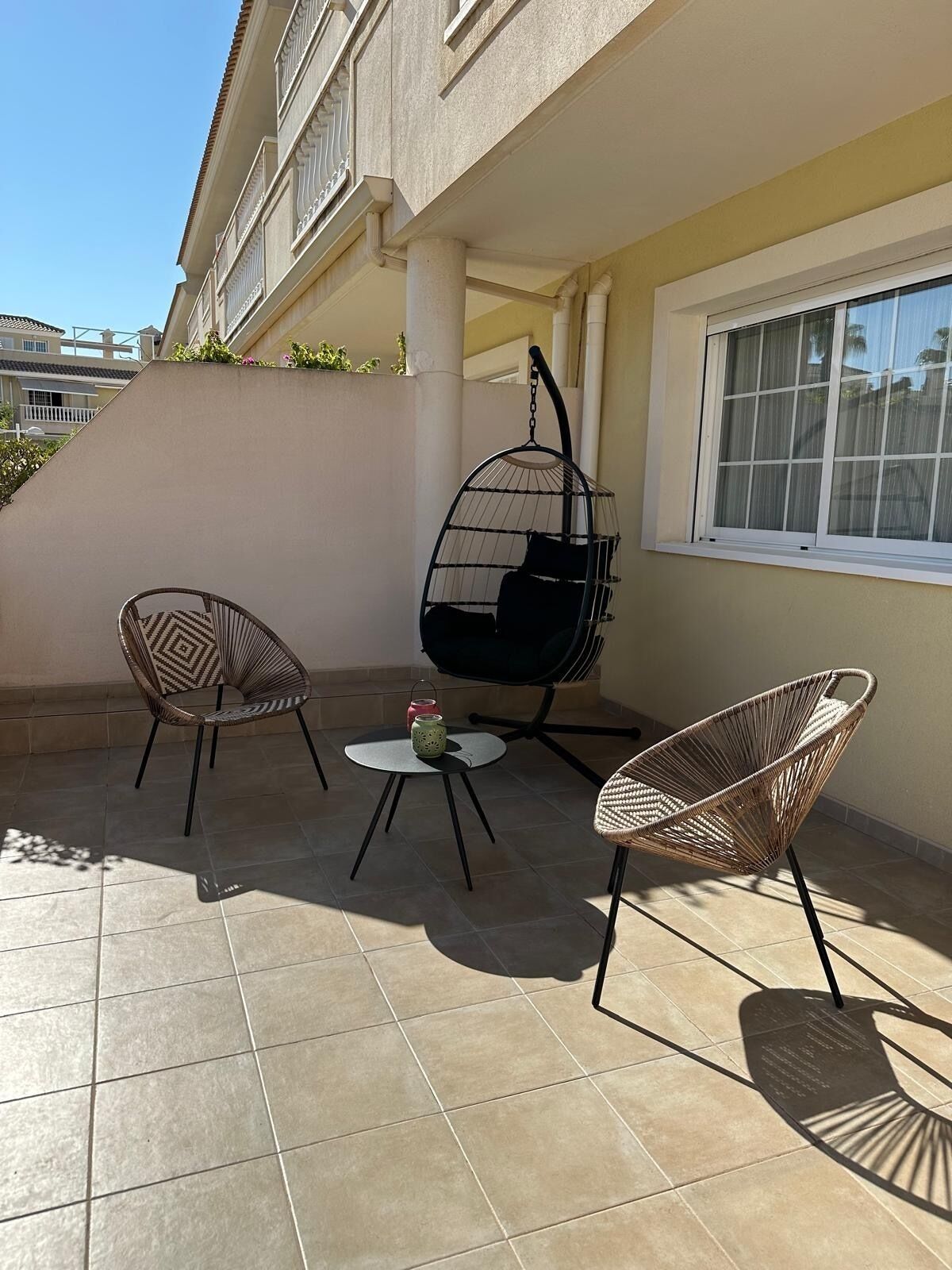 Une terrasse extérieure avec deux fauteuils en osier, un fauteuil suspendu et une petite table sur laquelle est posée une bougie.