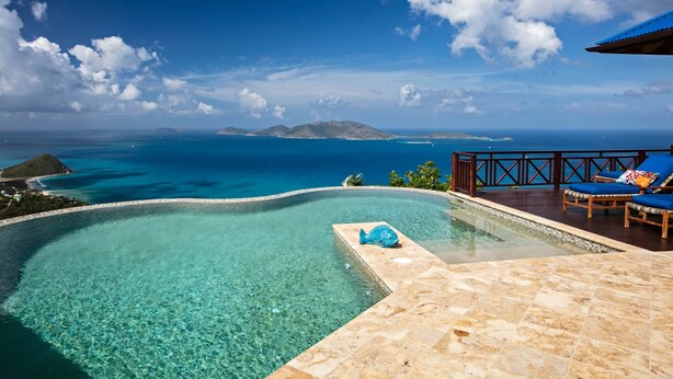 Uma piscina azul límpida com uma escultura de peixe, um deck de madeira com espreguiçadeiras e vista para o oceano e ilhas.