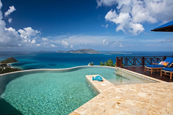 A clear blue pool with a fish sculpture, a wooden deck with lounge chairs, and a view of the ocean and islands.