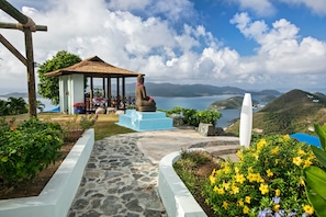 A gazebo with a stone statue, a stone pathway, and a view of the sea and hills.