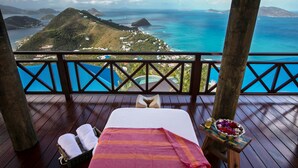 Um deck de madeira com uma cama, uma mesa pequena com uma tigela de frutas e vista para o oceano e as colinas.