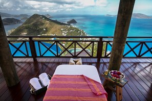 A wooden deck with a bed, a small table with a bowl of fruit, and a view of the ocean and hills.