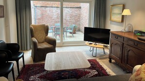 Un salon comprenant un canapé, un téléviseur, un meuble en bois et une vue sur un bâtiment en briques, visible à travers une porte vitrée.