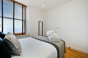 A bedroom with a bed, a window with blinds, a bedside table with towels, and a mirror.