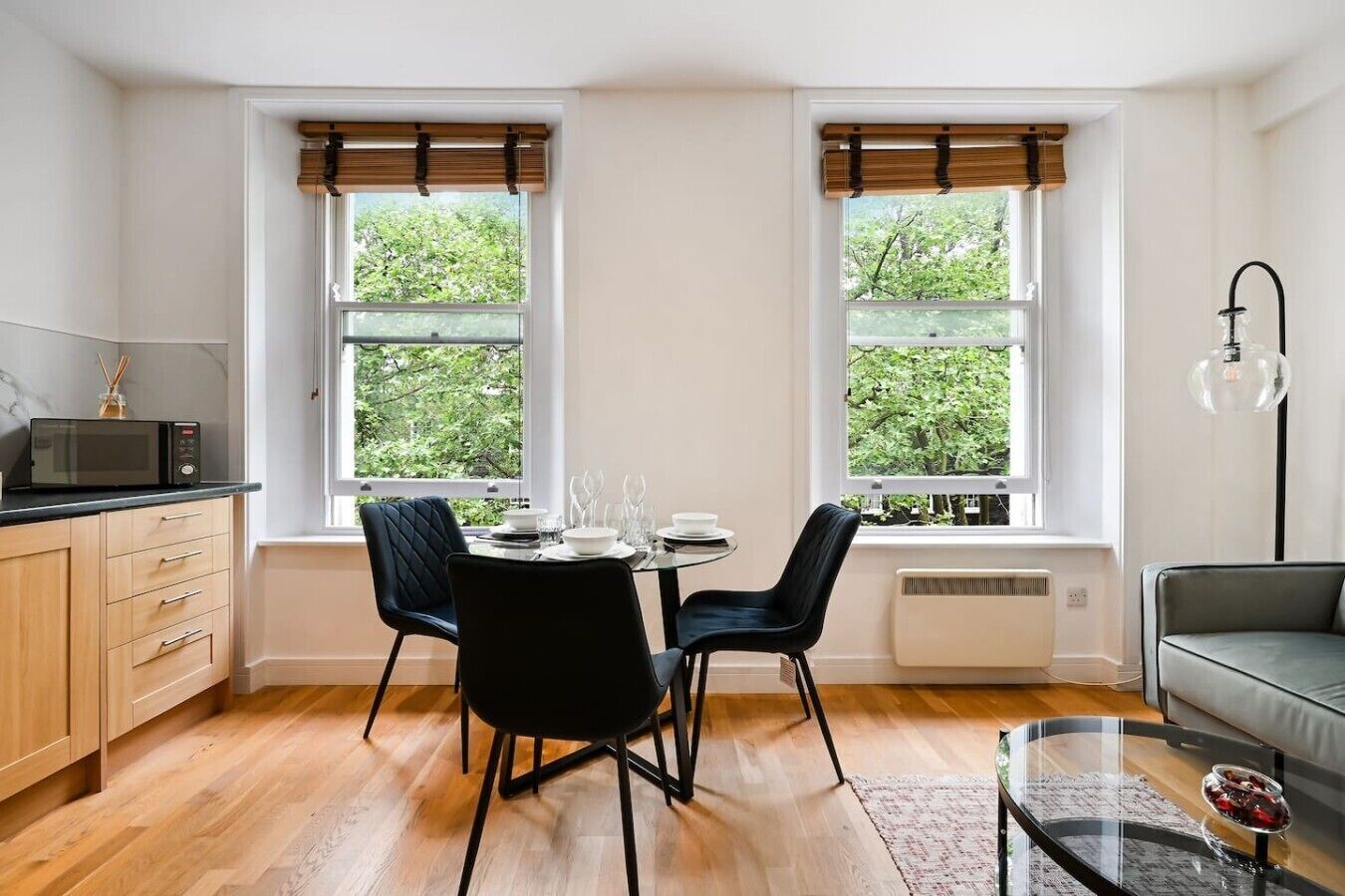 A modern kitchen with a dining table set for four, two windows with blinds, a microwave, and a sofa in a living area.