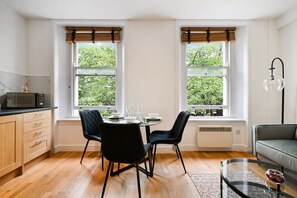 Une cuisine moderne avec une table à manger pour quatre personnes, deux fenêtres équipées de stores, un four à micro-ondes et un canapé dans l’espace salon.