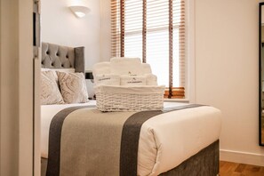 Ein ordentlich bezogenes Bett mit einer grau-weißen Streifenoptik, ein Korbwäscheschrank mit Handtüchern und ein grauer Kopfseitenverkleidung.
