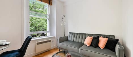 A living room with a grey sofa, black and peach cushions, a round glass coffee table, a black chair, and a window with a view of greenery.