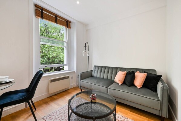A living room with a grey sofa, black and peach cushions, a round glass coffee table, a black chair, and a window with a view of greenery.
