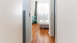 A narrow hallway leading to a bedroom with a bed, a chair, and a window with curtains.