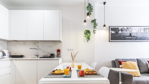 A modern kitchen with white cabinets, a dining table with chairs, and a grey sofa.