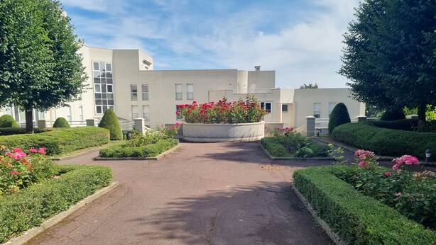 Un camino conduce a un edificio de diseño moderno, rodeado de setos cuidados y plantas con flores.