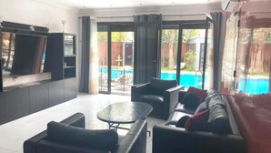 A modern living room with a pool view, featuring a sofa set, a TV, and a glass door leading to the pool. - Splendide Villa, Marrakech (Marrakech)