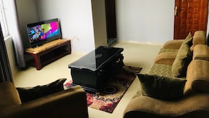 A living room with a TV on a wooden stand, a black suitcase on a rug, and a brown sofa. - SIMPLE GUEST HOUSE (Addis Ababa)