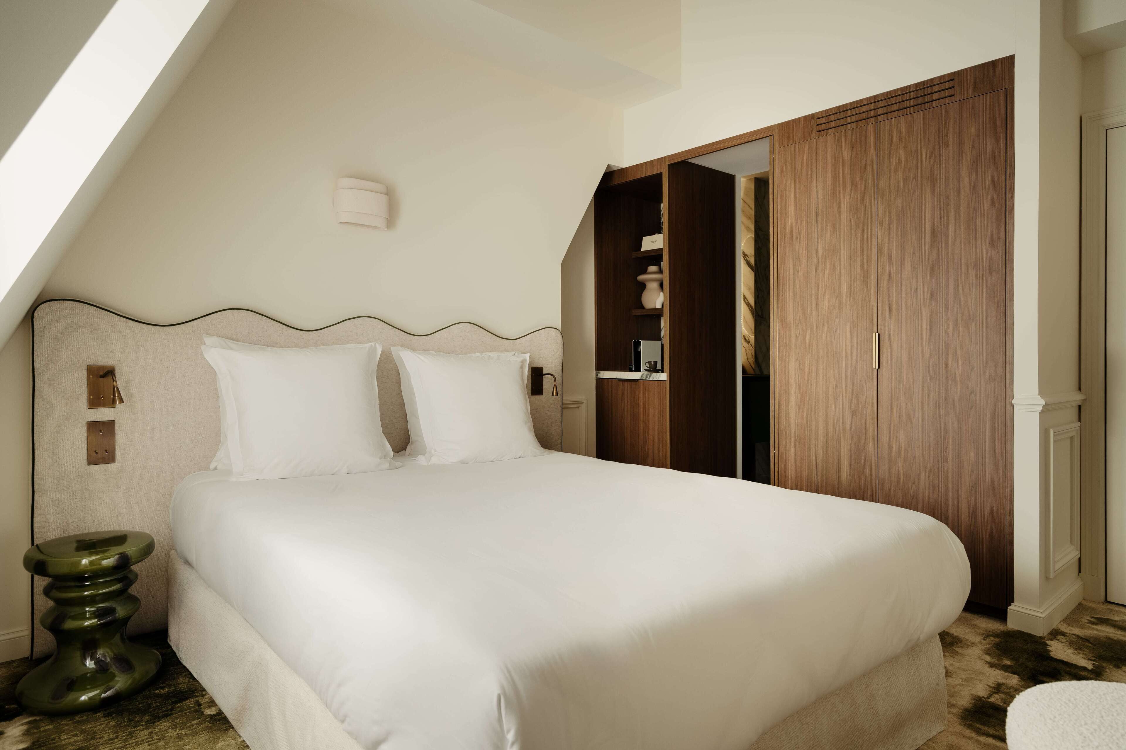 A neatly made bed in a room with a sloped ceiling, a wooden wardrobe, and a small green side table.