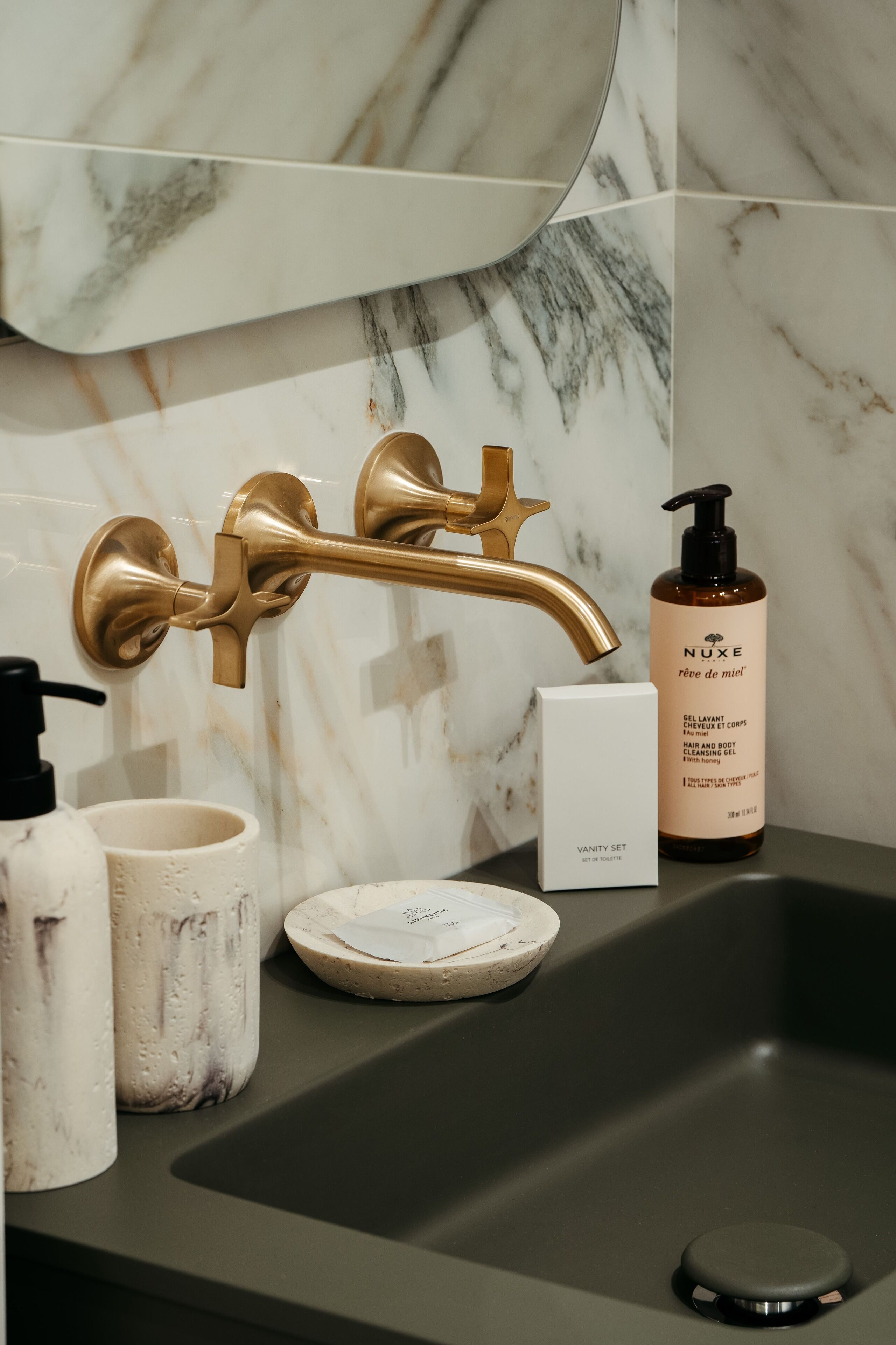 A bathroom vanity with a gold faucet, a Nuxe bottle, and a vanity set box.