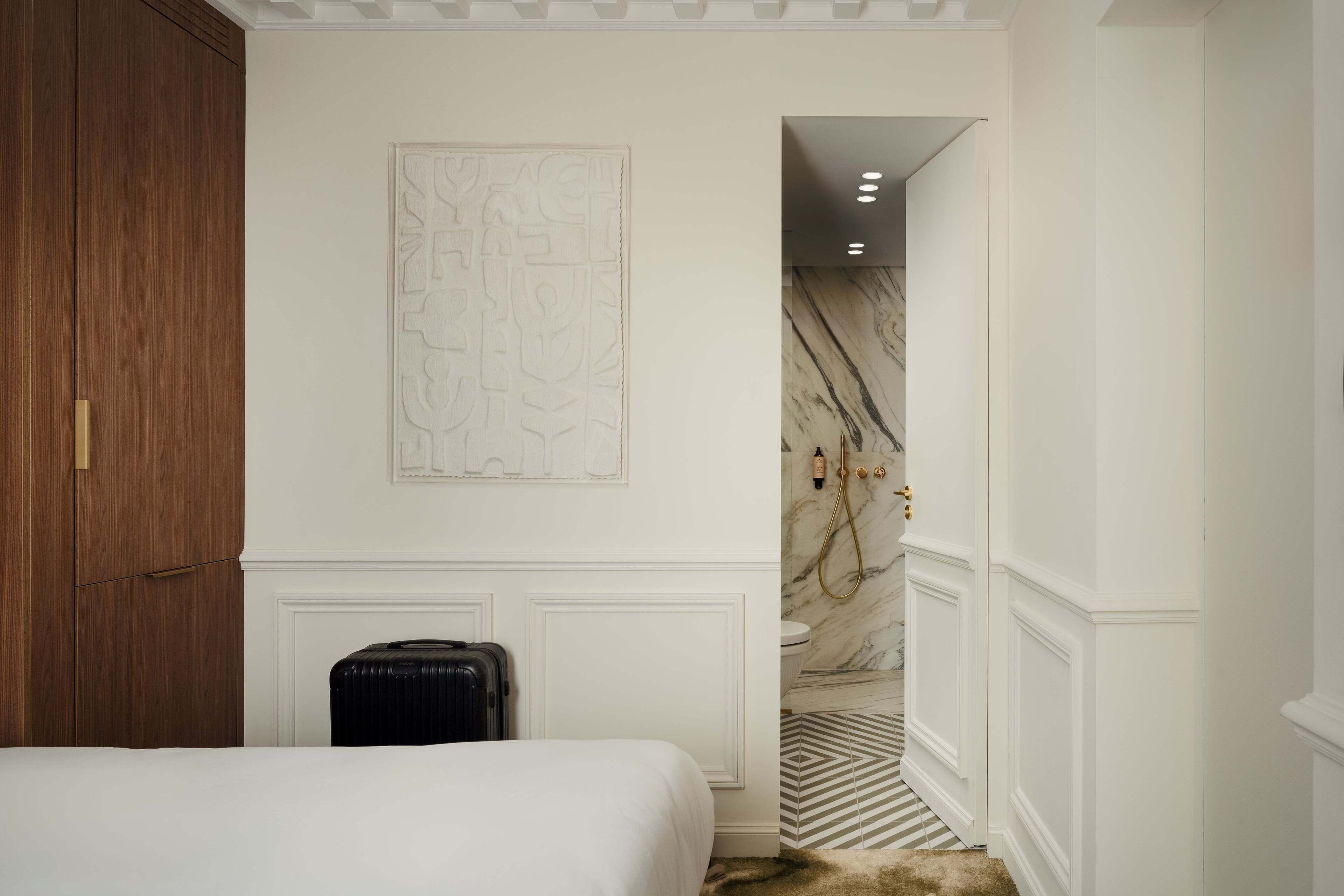 A hotel room with a bed, a suitcase, a marble wall, and a bathroom with a shower.