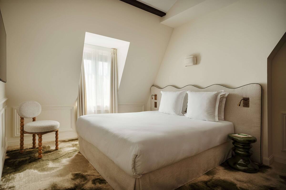 A bedroom with a large bed, a chair, a bedside table, and a window with curtains.