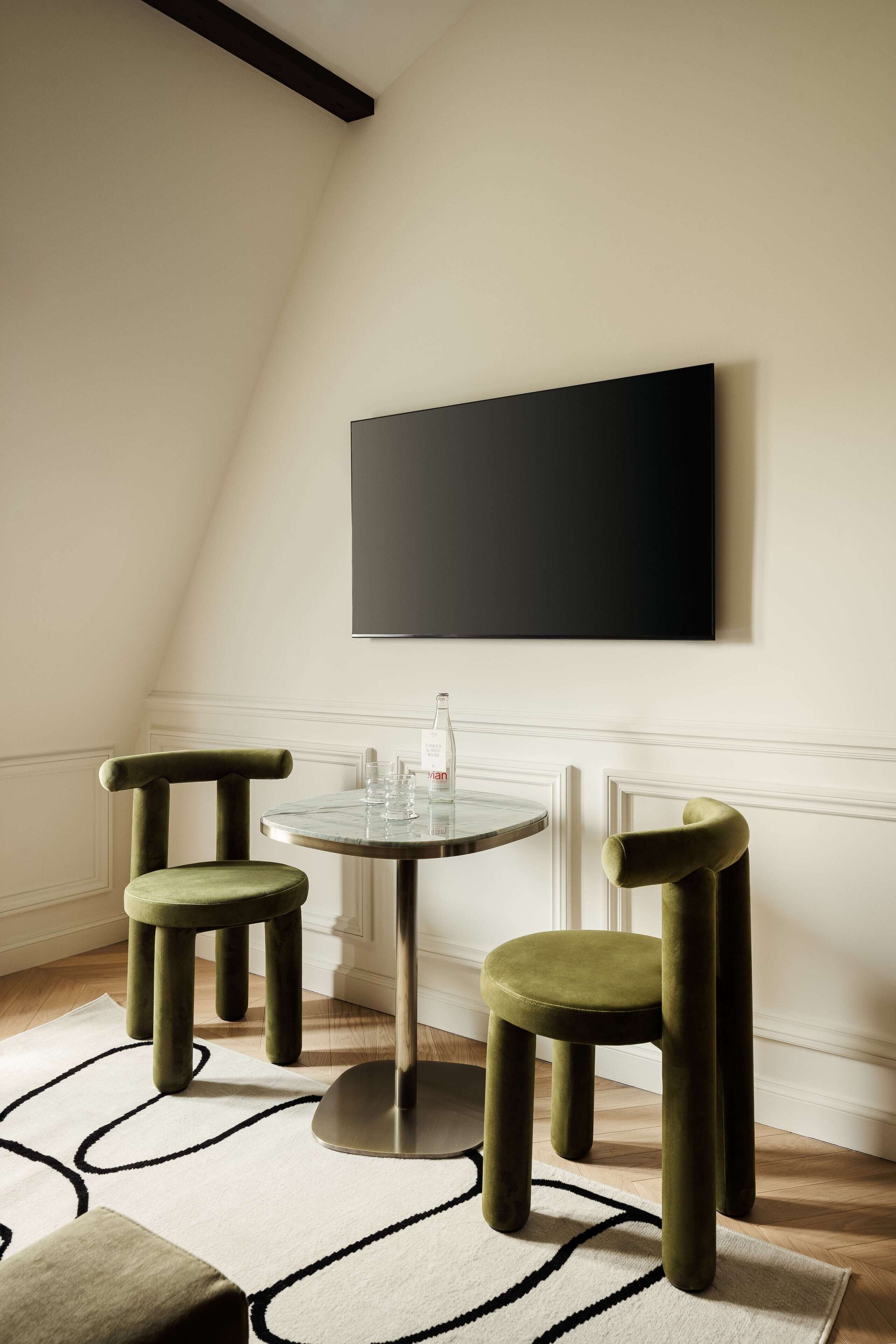 A dining area with a round table, two green chairs, a black wall-mounted TV, and a patterned rug.